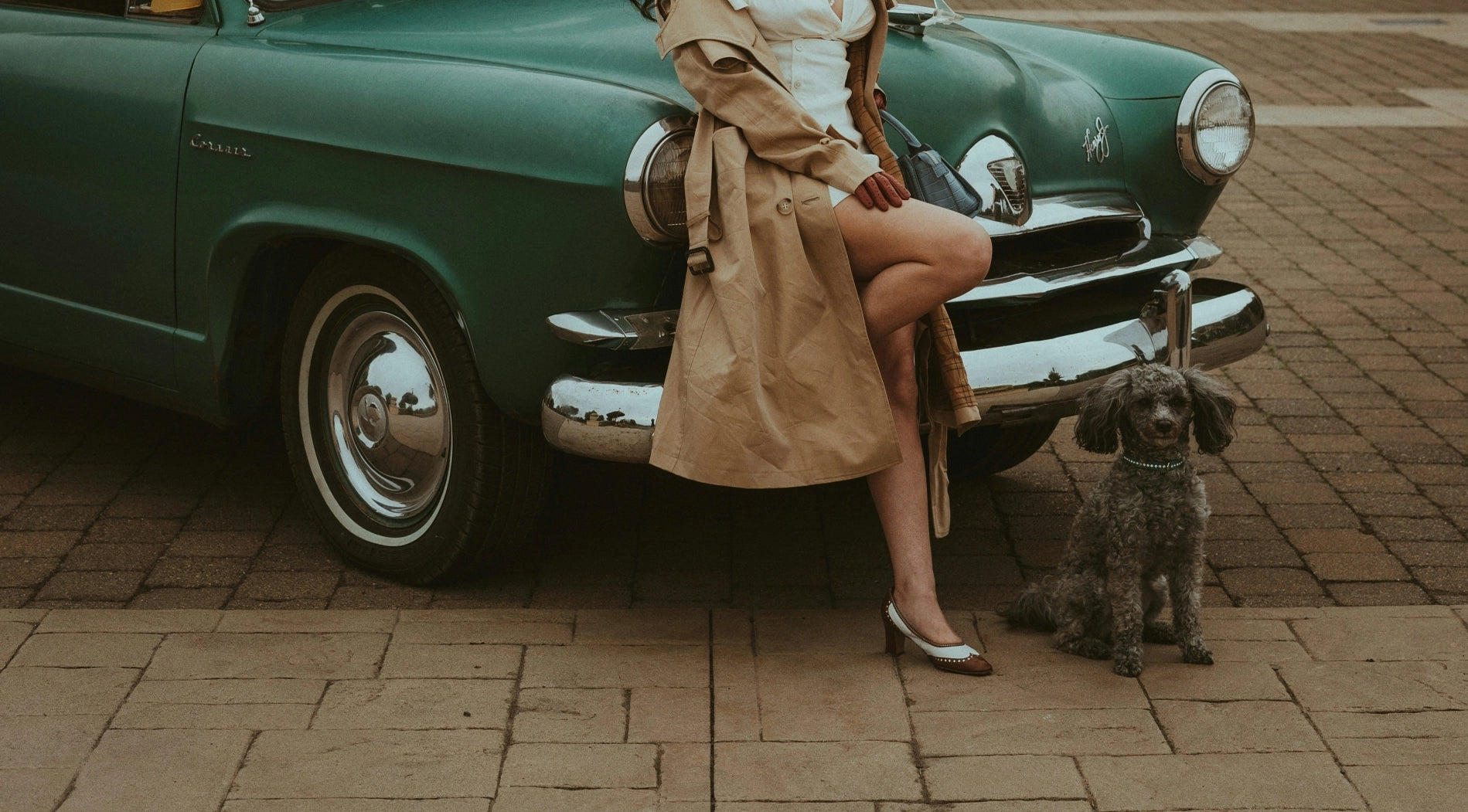 A woman sitting on the hood of a green car next to a dog.
